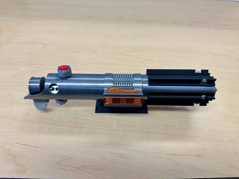 Puede incluir: Una r&eacute;plica de sable l&aacute;ser plateado con un mango negro y un detalle de color cobre. El sable l&aacute;ser est&aacute; montado en un soporte negro.