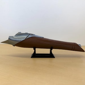 May include: A brown and silver-colored object, possibly a decorative item or prop, is displayed on a black stand. The object has a wooden-like texture on the main body and metallic accents. The design is elongated and aerodynamic.