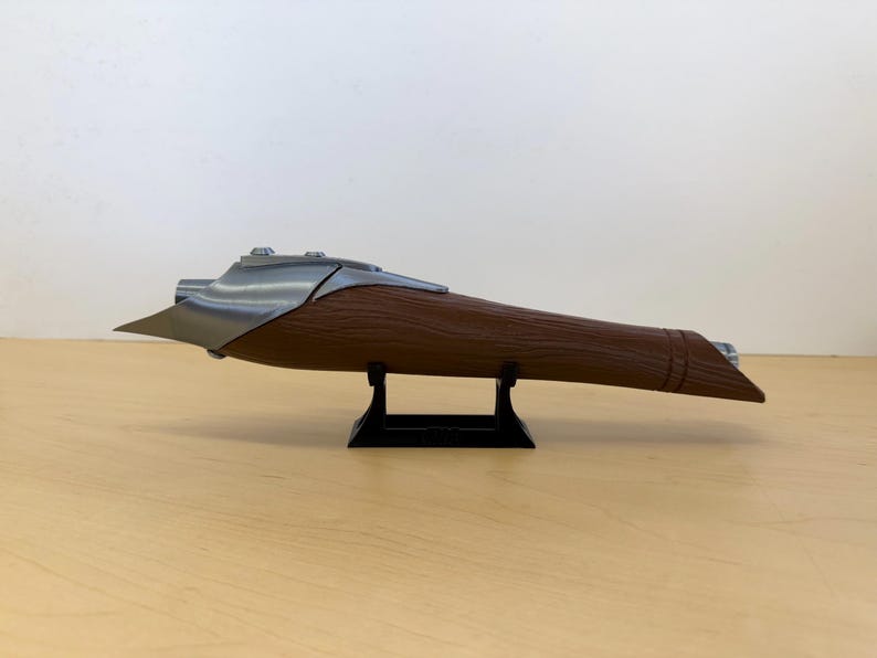 May include: A brown and silver-colored object, possibly a decorative item or prop, is displayed on a black stand. The object has a wooden-like texture on the main body and metallic accents. The design is elongated and aerodynamic.