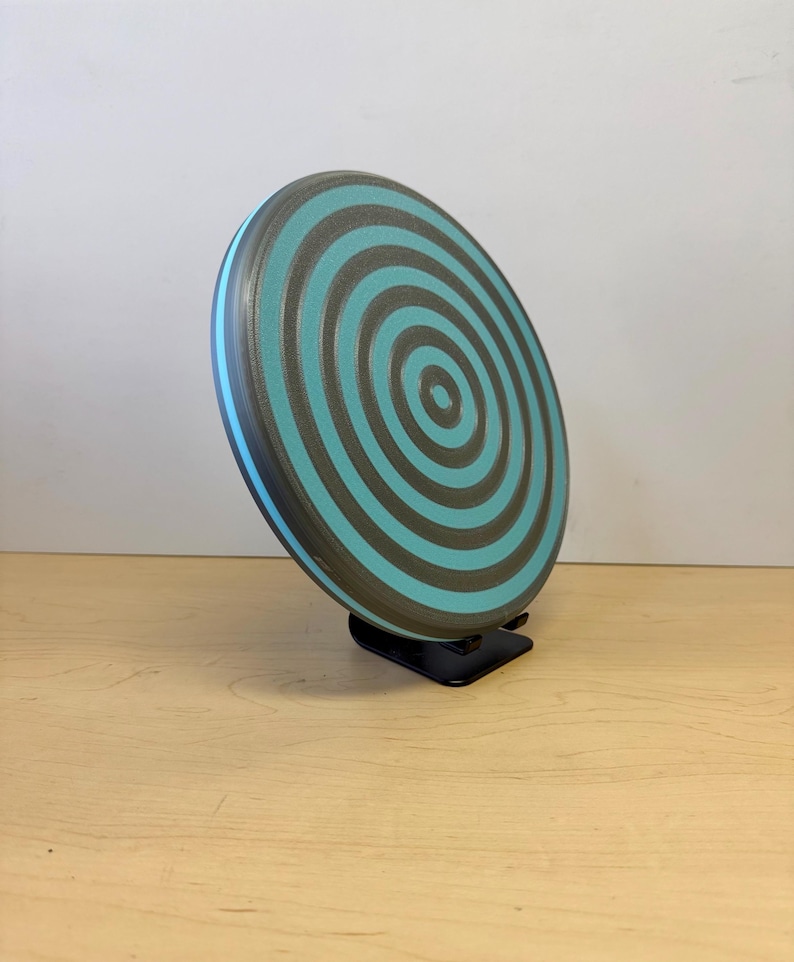May include: A teal and gray disc golf disc with a concentric circle design. The disc has a blue rim and is displayed on a black stand. The disc is made of a durable plastic material and is designed for outdoor sports.