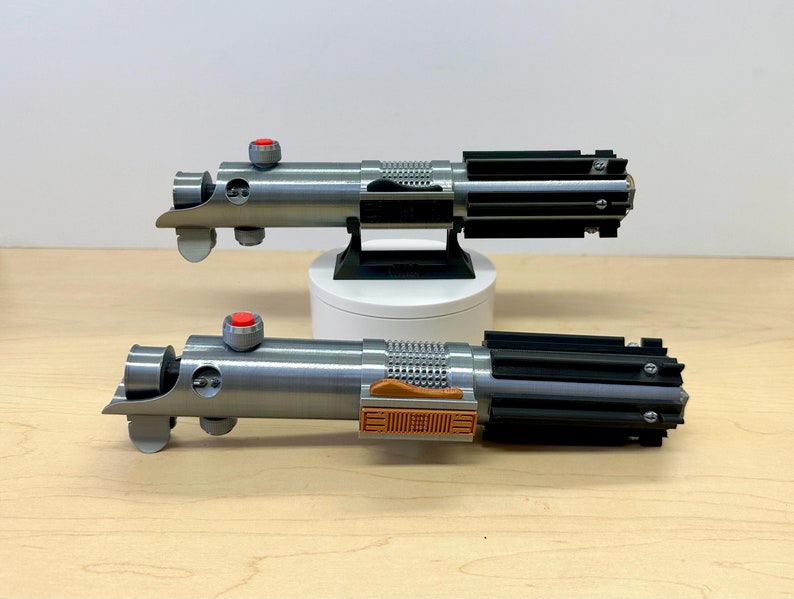 Puede incluir: Dos accesorios de sable de luz plateados y negros con botones rojos y un acento marr&oacute;n. Los sables de luz est&aacute;n sobre una superficie de madera.