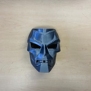 May include: A black and gray plastic mask with a detailed design resembling a face. The mask has a textured surface and a metallic sheen.