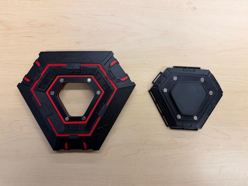 May include: Two black and red geometric objects with a hexagonal shape. The larger object has a central opening and red accents. The smaller object is solid black. Both have metallic accents.