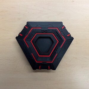 May include: A black and red hexagonal object with a recessed center. The object has a geometric design with red accents and a textured surface. The overall design is modern and industrial.