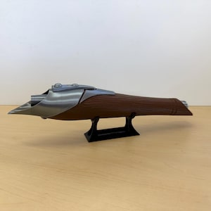 May include: A brown and silver prop weapon, possibly a futuristic blaster or rifle, is displayed on a black stand. The weapon has a wooden-like body with metallic accents and a unique, angular design. The prop is likely for cosplay or display.