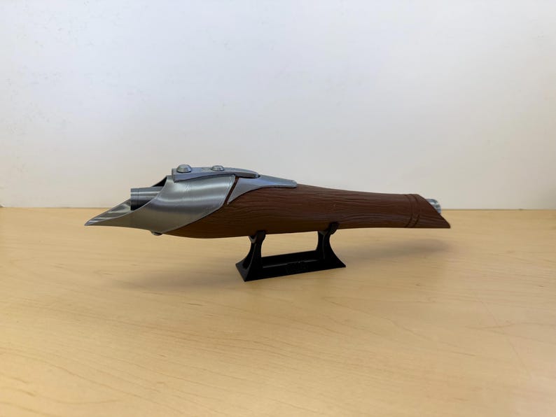 May include: A brown and silver prop weapon, possibly a futuristic blaster or rifle, is displayed on a black stand. The weapon has a wooden-like body with metallic accents and a unique, angular design. The prop is likely for cosplay or display.