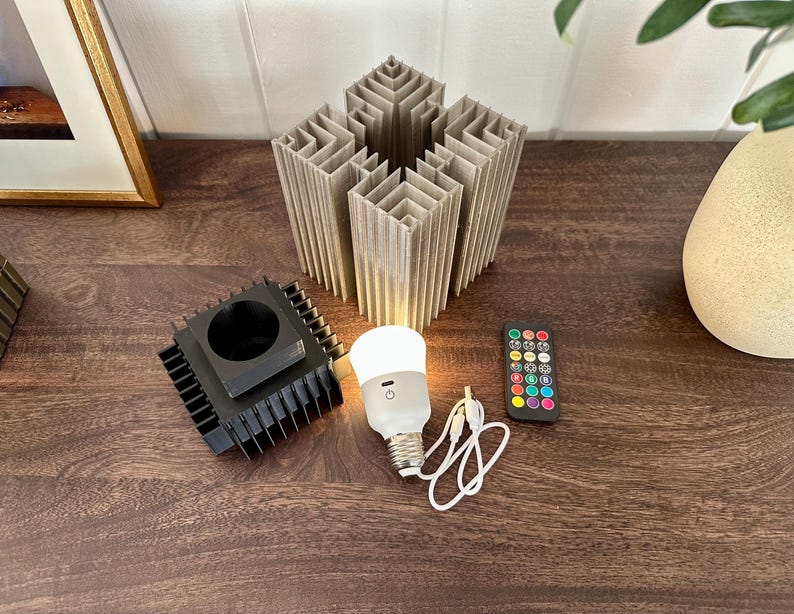 May include: A white light bulb with a black base and a remote control with coloured buttons. The light bulb is connected to a black metal base with a square shape. The remote control is on a wooden surface.