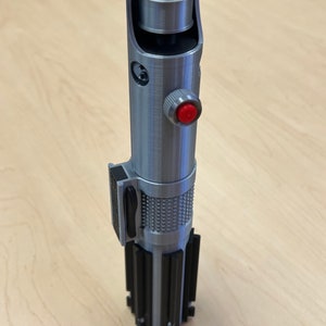 Puede incluir: Un accesorio de sable de luz plateado con un mango negro y un bot&oacute;n rojo. El sable de luz tiene un dise&ntilde;o detallado y una superficie texturizada.