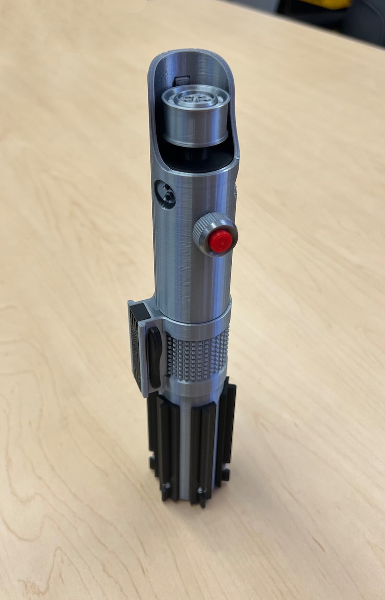 Puede incluir: Un accesorio de sable de luz plateado con un mango negro y un bot&oacute;n rojo. El sable de luz tiene un dise&ntilde;o detallado y una superficie texturizada.