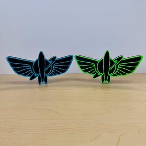 May include: Two standing emblems featuring a central rocket ship silhouette surrounded by stylized wings and a circular ring. One emblem has a bright blue outline against a black center, while the other has a vibrant neon green outline.