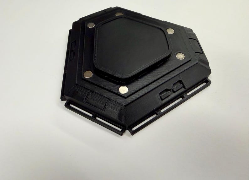 May include: Black, geometric-shaped object with a central, raised panel and several circular silver magnets. The object has a textured surface and appears to be made of a rigid material. The design includes rectangular cutouts and a layered construction.