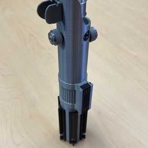 Puede incluir: Una empu&ntilde;adura de sable de luz plateada con detalles negros. La empu&ntilde;adura tiene una superficie texturizada y un peque&ntilde;o bot&oacute;n negro en el lateral.