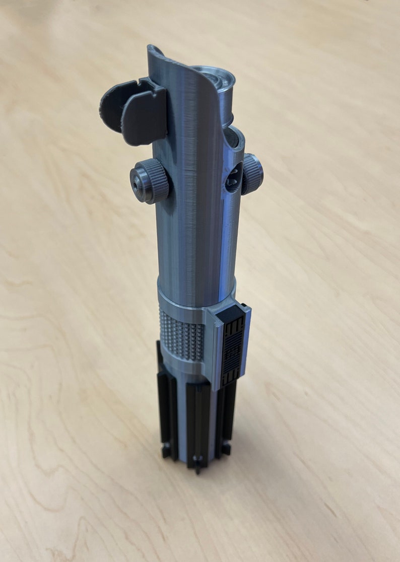 Puede incluir: Una empu&ntilde;adura de sable de luz plateada con detalles negros. La empu&ntilde;adura tiene una superficie texturizada y un peque&ntilde;o bot&oacute;n negro en el lateral.