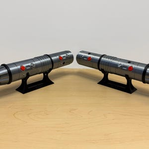 May include: Two metallic gray lightsabers with black accents and red buttons, displayed on black stands. The lightsabers have detailed designs and are positioned on a light brown wooden surface.
