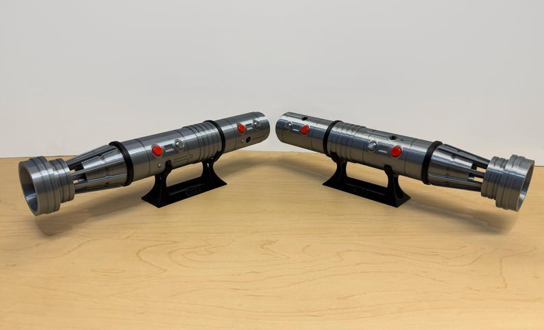 May include: Two metallic gray lightsabers with black accents and red buttons, displayed on black stands. The lightsabers have detailed designs and are positioned on a light brown wooden surface.