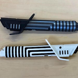 May include: Two toy lightsabers are displayed. The top one is black and gray with a silver handle. The bottom one is white and black. Both have a curved handle guard. The lightsabers are on a light brown surface.