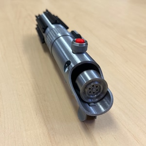 Puede incluir: Una r&eacute;plica de sable de luz plateado con un mango negro y un bot&oacute;n rojo. El sable de luz est&aacute; abierto, revelando el funcionamiento interno de la hoja.