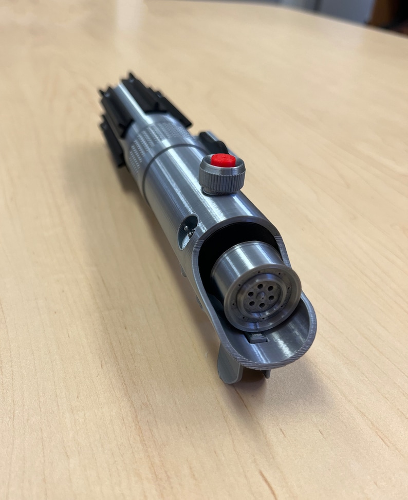 Puede incluir: Una r&eacute;plica de sable de luz plateado con un mango negro y un bot&oacute;n rojo. El sable de luz est&aacute; abierto, revelando el funcionamiento interno de la hoja.