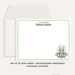 May include: A white rectangular note card with a thin green border, featuring the text a note from Kimberly Westin at the top and a small illustration of a green alien in a grey flying saucer in the bottom right corner, with a white envelope.