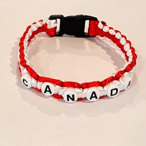 May include: A red, white, and black paracord bracelet. The bracelet features the word "CANADA" spelled out in white letter beads. The bracelet has a black plastic buckle.