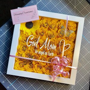 May include: A white frame filled with yellow paper roses and a pink bow. The text "God Mom An angel on Earth" is written in white on the roses.