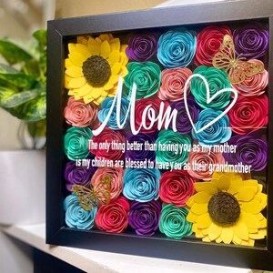 May include: A framed shadow box with a colorful paper flower arrangement. The flowers are in shades of pink, blue, purple, green, and yellow. The box also includes a gold butterfly and a heart with the word "Mom" written in white. The text below reads "The only thing better than having you as my mother is my children are blessed to have you as their grandmother."