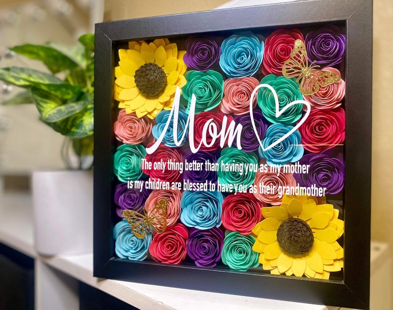 May include: A framed shadow box with a colorful paper flower arrangement. The flowers are in shades of pink, blue, purple, green, and yellow. The box also includes a gold butterfly and a heart with the word "Mom" written in white. The text below reads "The only thing better than having you as my mother is my children are blessed to have you as their grandmother."