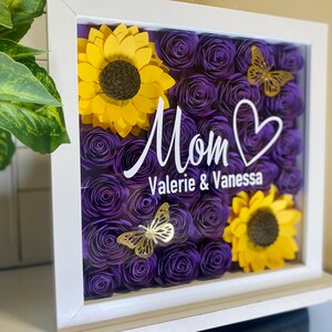 May include: A white frame with a glass front containing a purple and yellow paper flower arrangement. The flowers are arranged in a heart shape with a white inscription that reads "Mom Valerie & Vanessa".