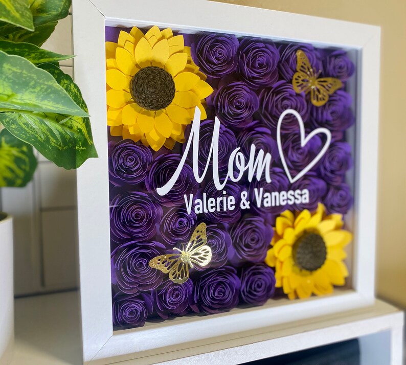 May include: A white frame with a glass front containing a purple and yellow paper flower arrangement. The flowers are arranged in a heart shape with a white inscription that reads "Mom Valerie & Vanessa".