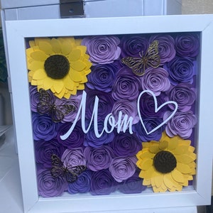 May include: A white frame with a glass front displays a purple and yellow paper flower arrangement. The flowers are arranged in a heart shape and the word "Mom" is written in white script across the center.