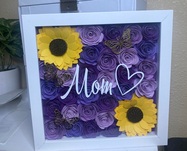 May include: A white frame with a glass front displays a purple and yellow paper flower arrangement. The flowers are arranged in a heart shape and the word "Mom" is written in white script across the center.