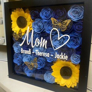 May include: A framed shadow box with blue and yellow paper flowers, butterflies, and the word "Mom" with the names "Brandi, Theresa, Jackie" written below.