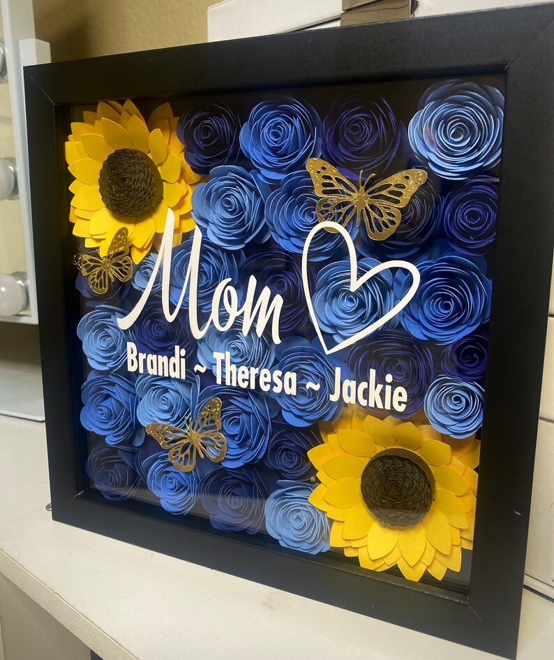 May include: A framed shadow box with blue and yellow paper flowers, butterflies, and the word "Mom" with the names "Brandi, Theresa, Jackie" written below.