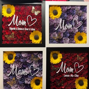 May include: Four framed shadow boxes with paper flowers. Each box has a different arrangement of red, purple, and yellow paper flowers. The boxes have white frames and black mats. The text "Mom" is written in white on each box, with different names below each.