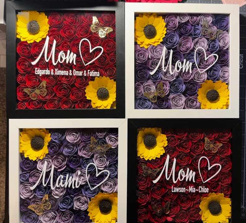 May include: Four framed shadow boxes with paper flowers. Each box has a different arrangement of red, purple, and yellow paper flowers. The boxes have white frames and black mats. The text "Mom" is written in white on each box, with different names below each.