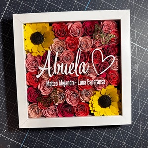 May include: A white frame with a heart and the word "Abuela" in white script. The frame is filled with paper flowers in shades of pink, red, and yellow. There are two butterflies in the frame, one gold and one brown.