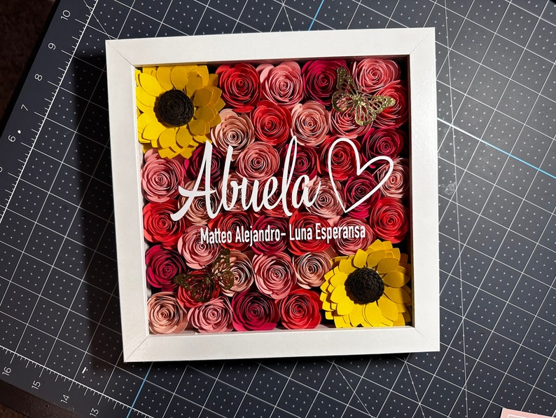 May include: A white frame with a heart and the word "Abuela" in white script. The frame is filled with paper flowers in shades of pink, red, and yellow. There are two butterflies in the frame, one gold and one brown.
