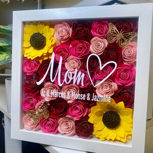 May include: A white frame filled with pink, red, and yellow paper flowers. The flowers are arranged in a heart shape with a white heart drawn in the center. The text "Mom" is written in white cursive script with the names "Jc & Marcos & Monse & Jazmine" written below.