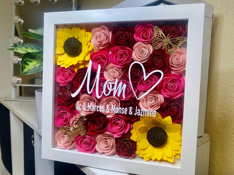 May include: A white frame filled with pink, red, and yellow paper flowers. The flowers are arranged in a heart shape with a white heart drawn in the center. The text "Mom" is written in white cursive script with the names "Jc & Marcos & Monse & Jazmine" written below.