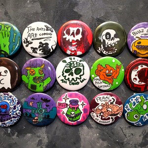 May include: A set of 12 round pins with various colorful designs and text. The pins feature a variety of themes, including spooky, funny, and pop culture references. Some of the text on the pins includes "The Rats Are Coming", "I Am Dead Lol", "Hail Fish Satan", and "Bugz 4 Lyf".
