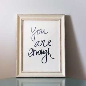 May include: A white framed print with the text "you are enough" written in black cursive script. The print is sitting on a light blue table with wooden legs.