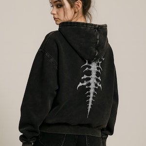 Op de afbeelding: Zwarte hoodie met een verweerde uitstraling, met een witte ruggengraat op de rug. De hoodie heeft een capuchon en lange mouwen. Het model draagt een zwarte short.