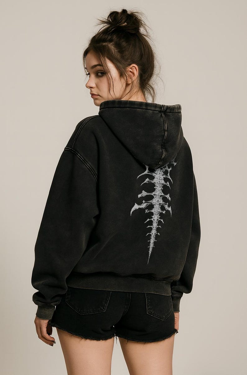 Op de afbeelding: Zwarte hoodie met een verweerde uitstraling, met een witte ruggengraat op de rug. De hoodie heeft een capuchon en lange mouwen. Het model draagt een zwarte short.