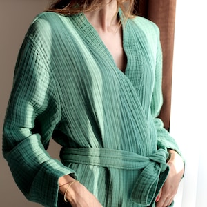 May include: A sage green robe made of textured muslin fabric featuring long sleeves, a shawl collar, and a matching tie belt at the waist. The garment includes two front patch pockets and is designed with a relaxed fit and a soft, crinkled appearance.