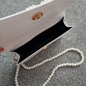 May include: White faux leather handbag with a gold clasp and a pearl chain strap.