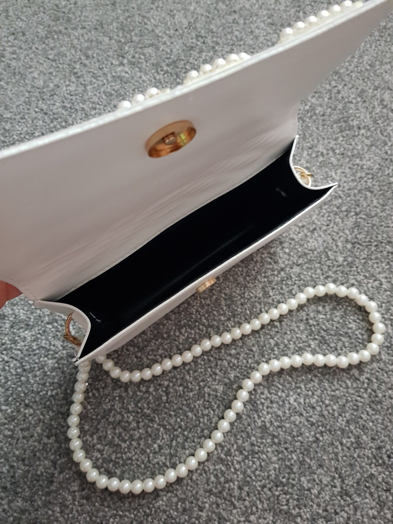 May include: White faux leather handbag with a gold clasp and a pearl chain strap.