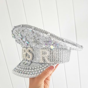 Op de afbeelding: Zilveren pailletten hoed met de letters "RSVR" in sprankelende versieringen. De hoed heeft een klep en is versierd met kristallen en parels. De hoed is een decoratief accessoire.