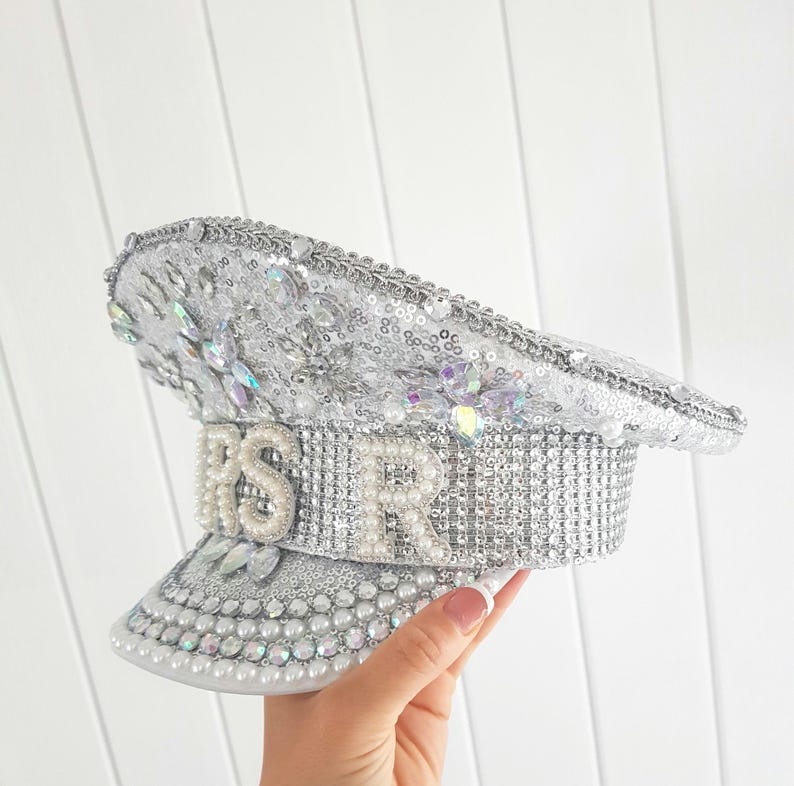 Op de afbeelding: Zilveren pailletten hoed met de letters "RSVR" in sprankelende versieringen. De hoed heeft een klep en is versierd met kristallen en parels. De hoed is een decoratief accessoire.