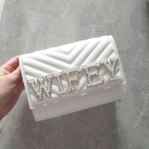 May include: White quilted clutch purse with the word "WIFEY" spelled out in silver letters with pearl accents.