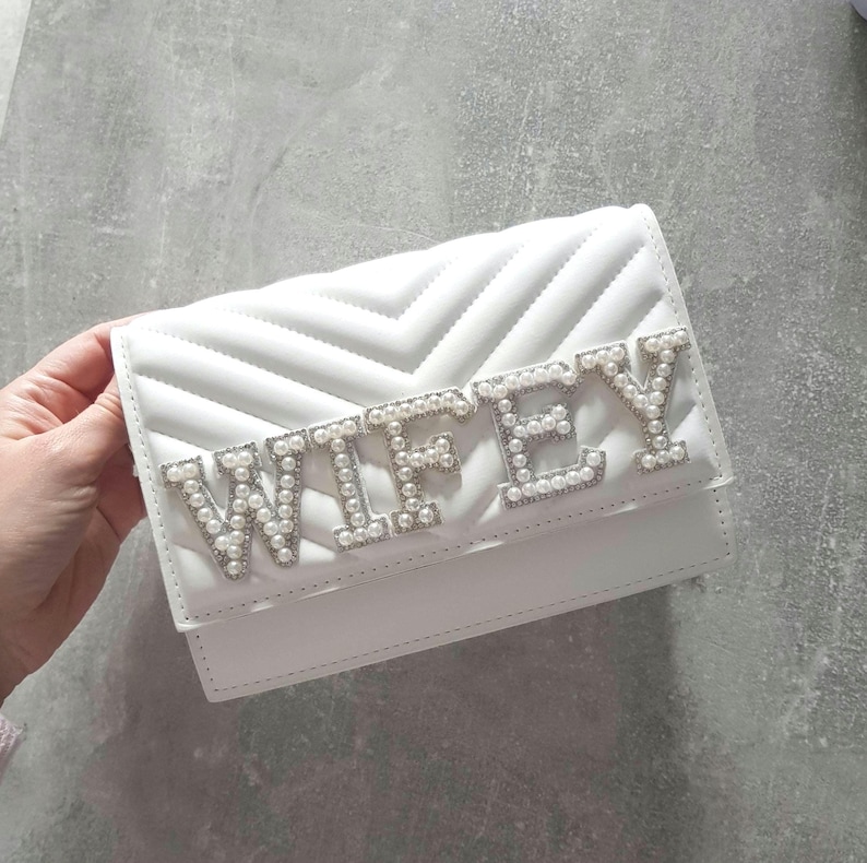May include: White quilted clutch purse with the word "WIFEY" spelled out in silver letters with pearl accents.
